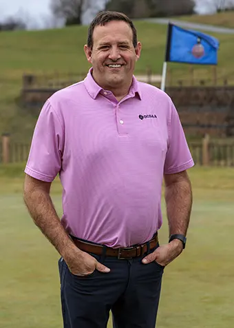 Man in a pink polo shirt standing on a golf course green with a blue flag in the background, smiling with hands in pockets.