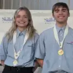 Two smiling SkillsUSA competitors wearing blue shirts with gold medals on stage beside a first-place sign.