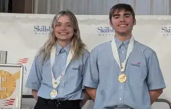 Two smiling SkillsUSA competitors wearing blue shirts with gold medals on stage beside a first-place sign.