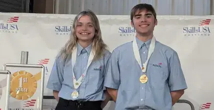 Two smiling SkillsUSA competitors wearing blue shirts with gold medals on stage beside a first-place sign.