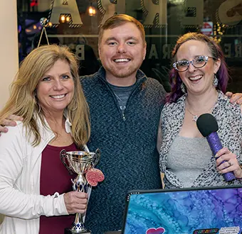 Three smiling people posing at an event: a woman on the left holding a trophy, a man in the middle, and a woman on the right with a microphone and laptop visible.
