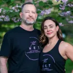 Two adults in black The MET shirts posing outdoors under flowering pink blossoms, smiling at the camera.