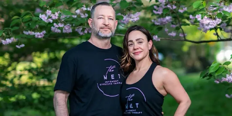 Two adults in black The MET shirts posing outdoors under flowering pink blossoms, smiling at the camera.