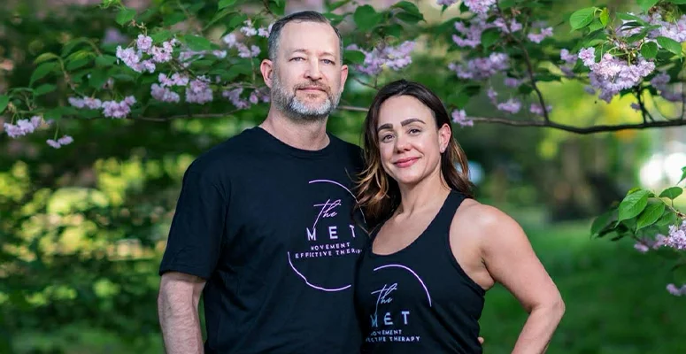 Two adults in black The MET shirts posing outdoors under flowering pink blossoms, smiling at the camera.