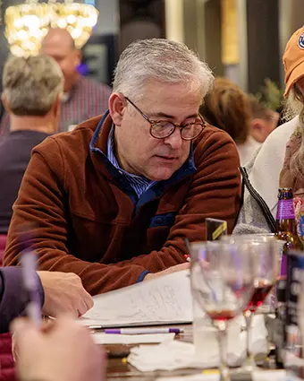 Man with glasses in a brown jacket reads documents at a busy dinner table, wine glasses nearby.