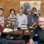 Group of seven adults smiling and seated around a wooden table in a brick-walled sports bar, Ravens logos on the wall, drinks and condiments on the table.