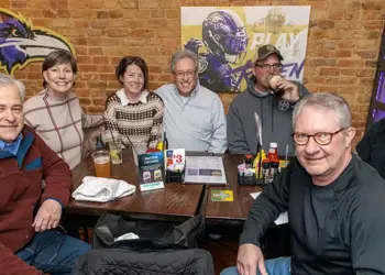 Group of seven adults smiling and seated around a wooden table in a brick-walled sports bar, Ravens logos on the wall, drinks and condiments on the table.