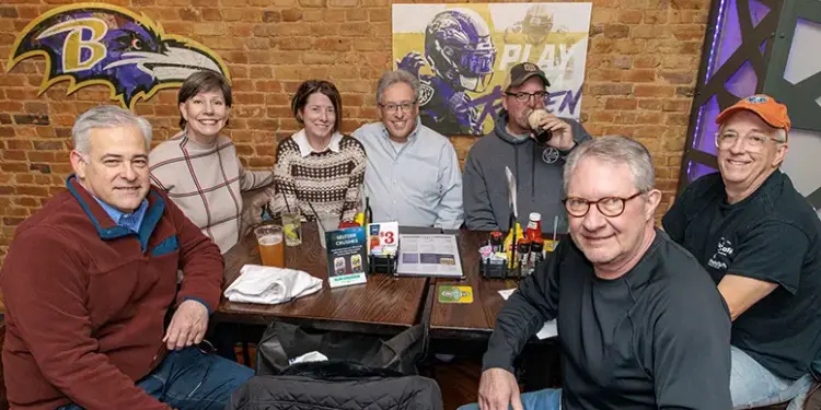 Group of seven adults smiling and seated around a wooden table in a brick-walled sports bar, Ravens logos on the wall, drinks and condiments on the table.