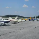 Line of small propeller airplanes parked in a row on a sunny airport tarmac under a blue sky
