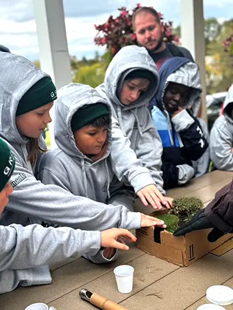 Group of kids in gray hoodies gathered around a wooden box, planting or inspecting soil, under adult supervision outdoors.