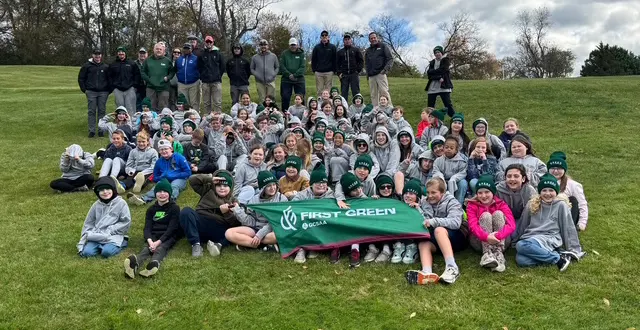 Large group of students and instructors posing on a grassy hill, holding a green First Green banner.