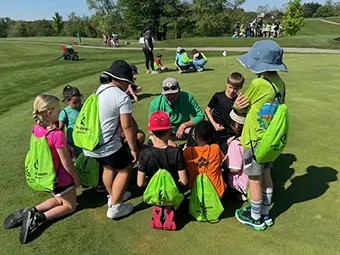 Group of children sitting in a circle on a grassy field with bright green backpacks, listening to instructors at an outdoor camp.