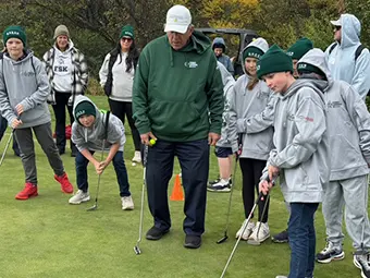 Youth golf lesson: coach demonstrates putting to a group on a practice green outside in fall colors organic setting.