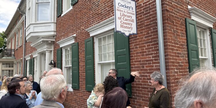 Crowd of people gathered outside a brick building with a hanging sign reading 'Cockey's Library of Carroll County History' outside the Historical Society.
