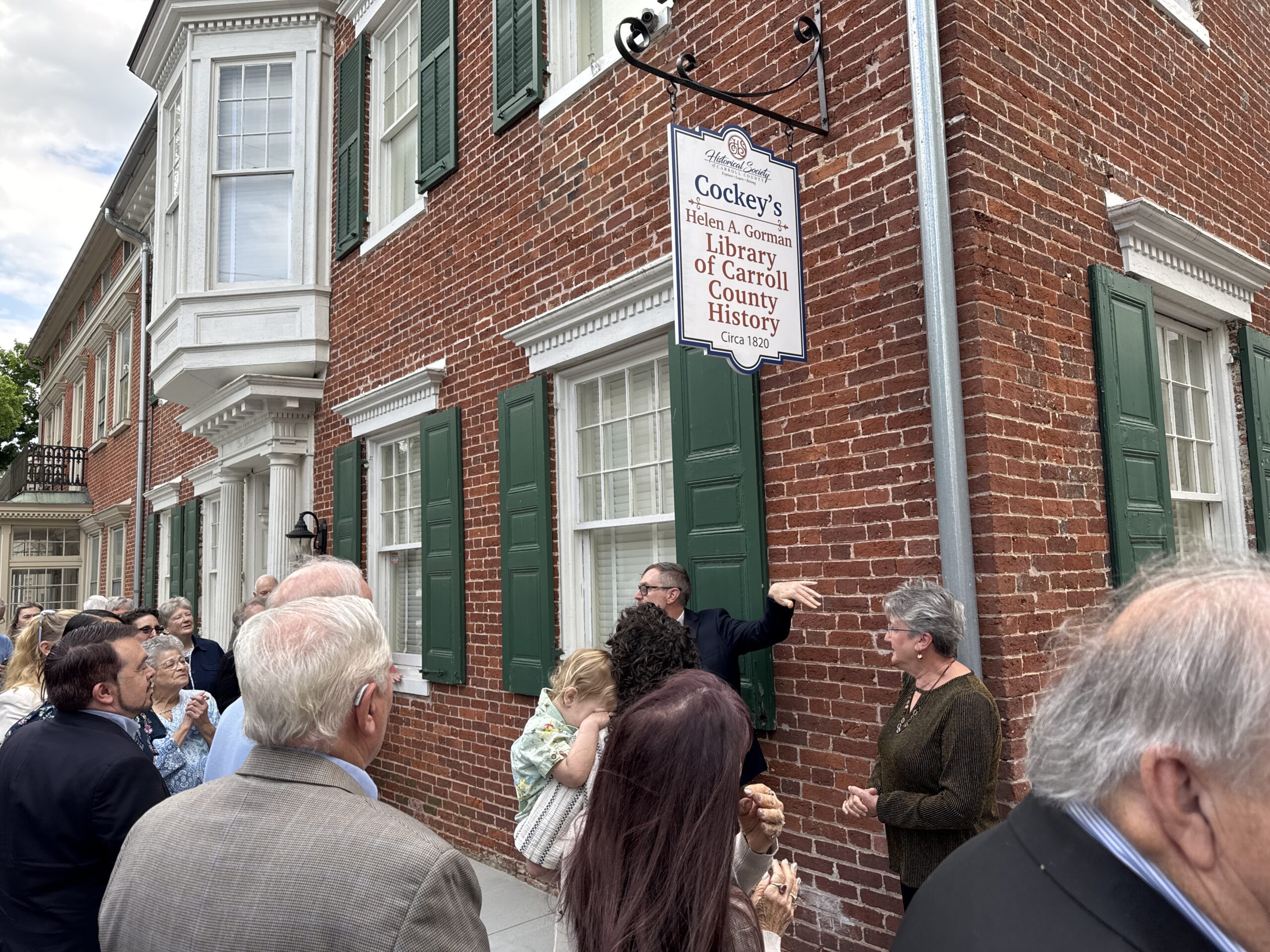 Crowd of people gathered outside a brick building with a hanging sign reading 'Cockey's Library of Carroll County History' outside the Historical Society.