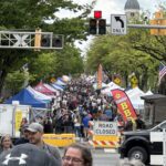 Crowded street festival with vendor tents, American flags, and signs reading ROAD CLOSED along a tree-lined avenue.