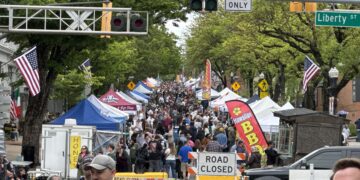 Crowded street festival with vendor tents, American flags, and signs reading ROAD CLOSED along a tree-lined avenue.