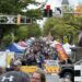 Crowded street festival with vendor tents, American flags, and signs reading ROAD CLOSED along a tree-lined avenue.