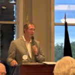 Man in a gray blazer speaks into a handheld microphone at a wooden podium during a meeting, with audience heads visible in the foreground and large windows showing a blue sky outside.