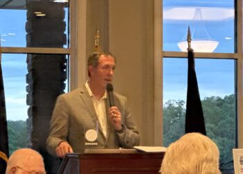 Man in a gray blazer speaks into a handheld microphone at a wooden podium during a meeting, with audience heads visible in the foreground and large windows showing a blue sky outside.