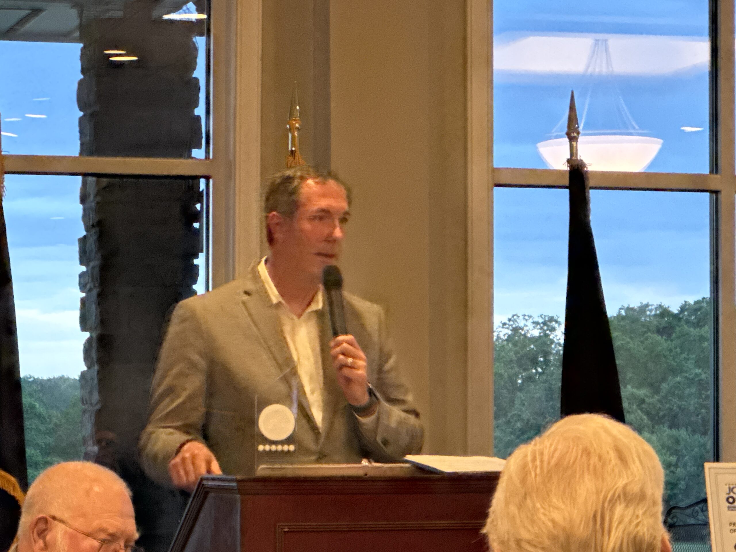 Man in a gray blazer speaks into a handheld microphone at a wooden podium during a meeting, with audience heads visible in the foreground and large windows showing a blue sky outside.