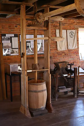 Historic wooden wine press on display in a brick-walled museum, with a wooden barrel beneath and old equipment in the background.
