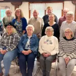 Group of fourteen seniors posing for a photo in a bright community room, with seven seated in front and seven standing behind, casual attire.