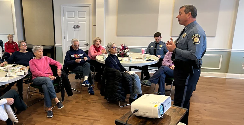 Senior adults seated at round tables in a community room, listening to a uniformed police officer giving a talk.