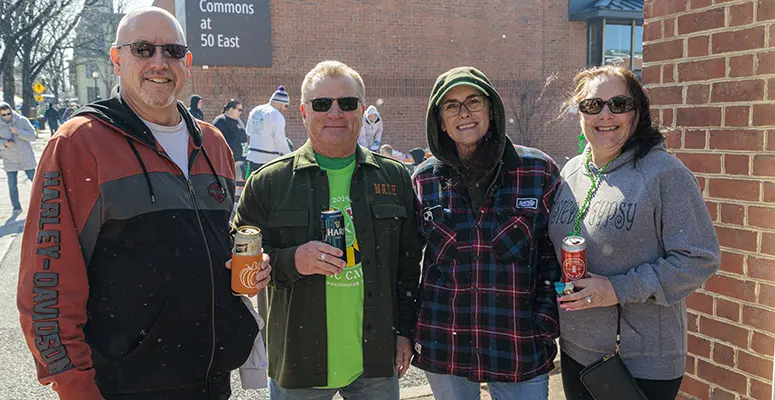 Four adults standing together outdoors at a community event, each holding a drink and smiling.