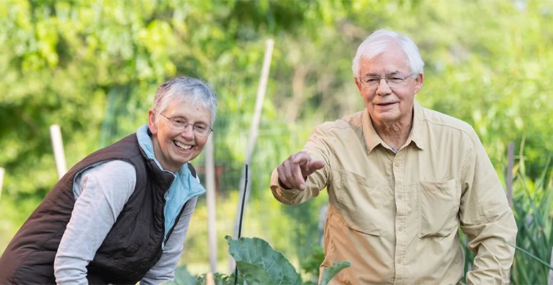 Two smiling seniors gardening together in a lush outdoor yard.