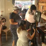 Group of people filming a video in a cafe, with a camera on a tripod and a monitor setup in the center of the scene.