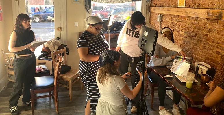 Group of people filming a video in a cafe, with a camera on a tripod and a monitor setup in the center of the scene.