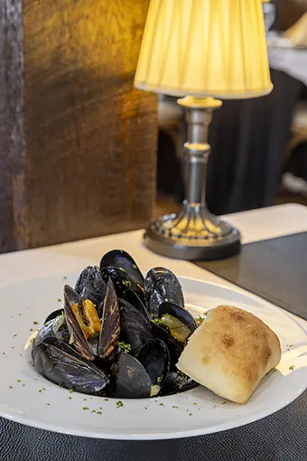 Plate of black mussels with a bread roll on a restaurant table, garnished with herbs and a warm lamp glow in the background.