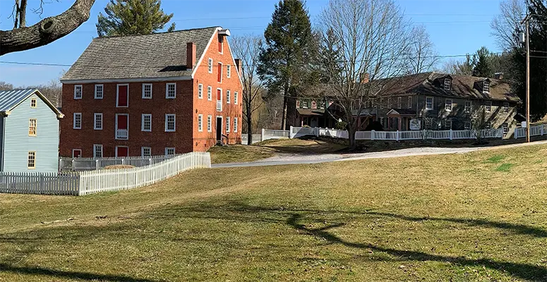 Brick building with a gabled roof and red doors along a street, white picket fences, and bare trees on a sunny day in a small town.