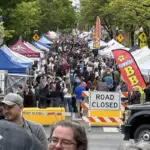 Crowded street festival with numerous vendor tents, American flags, and a road-closed barrier stretching along the avenue.