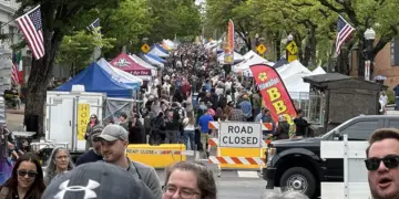 Crowded street festival with numerous vendor tents, American flags, and a road-closed barrier stretching along the avenue.