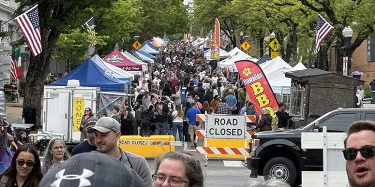Crowded street festival with numerous vendor tents, American flags, and a road-closed barrier stretching along the avenue.