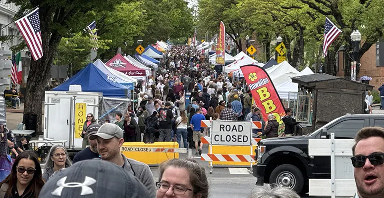 Crowded street festival with numerous vendor tents, American flags, and a road-closed barrier stretching along the avenue.