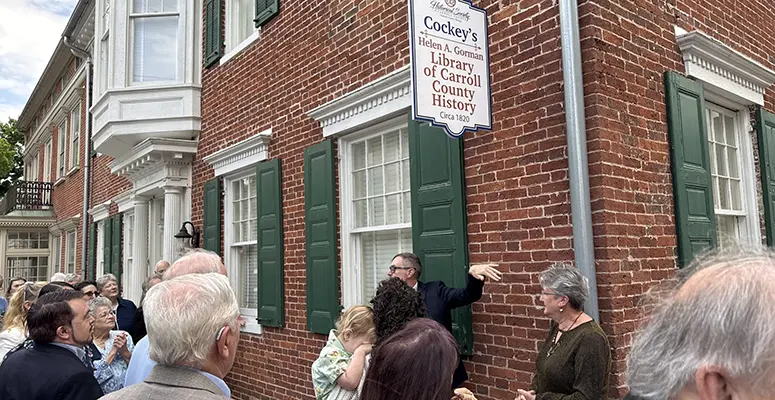 Crowd gathers outside a brick historic library building with green shutters and a hanging sign for Cockey's Helen B. Gorman Library of Carroll County History.