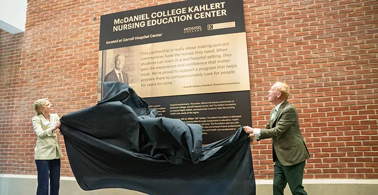 Two people unveil a plaque for the McDaniel College Kahler Nursing Education Center against a brick wall outside a hospital setting.