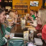 Two women sit across from each other at a long communal table in a lively restaurant, toasting with drinks and smiling.