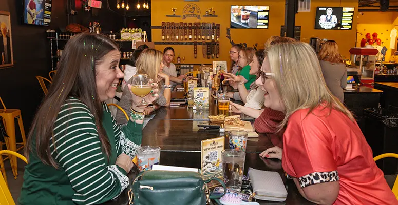 Two women sit across from each other at a long communal table in a lively restaurant, toasting with drinks and smiling.