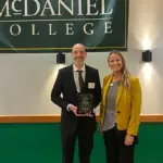 Two adults stand on a stage under a McDaniel College banner; man in a suit holds an award plaque, woman in a yellow blazer smiles beside him.