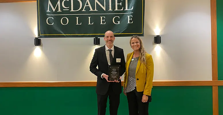 Two adults stand on a stage under a McDaniel College banner; man in a suit holds an award plaque, woman in a yellow blazer smiles beside him.