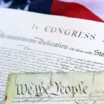 Close-up of the U.S. Declaration of Independence and a parchment reading 'We the People,' with an American flag in the background.