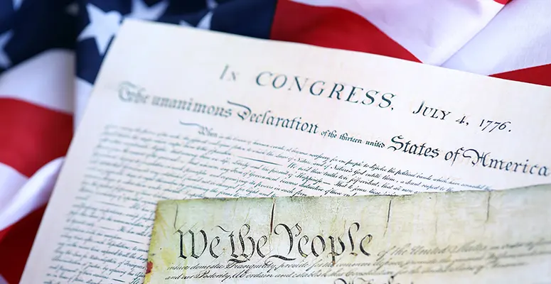 Close-up of the U.S. Declaration of Independence and a parchment reading 'We the People,' with an American flag in the background.