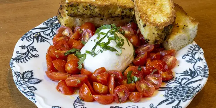 Burrata Pomodoro_1 - Carroll News | Carroll Publications | Carroll County, MD Plate of chopped cherry tomatoes around a burrata ball, topped with basil, with toasted bread slices on a blue-and-white patterned plate.