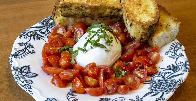 Plate of chopped cherry tomatoes around a burrata ball, topped with basil, with toasted bread slices on a blue-and-white patterned plate.