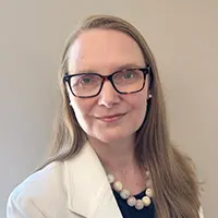 Headshot of a professional woman with glasses, shoulder-length hair, wearing a white blazer and pearl necklace against a neutral background.