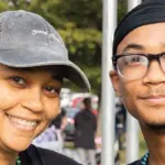 Two smiling people posing together at an outdoor event, wearing bead necklaces and casual clothing.